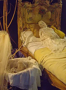 Human sculpture of woman in yellow bed with cradle in foreground.
