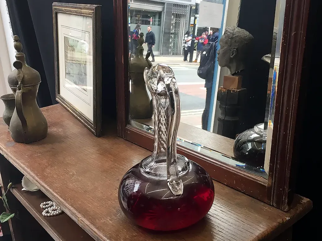 Book shelf with picture frame, a glass flagon with red liquid and a mirror partly reflecting a real life street scene.