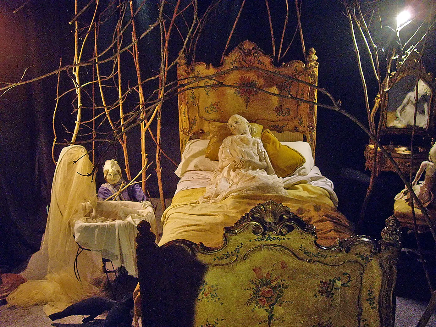 Antique bedroom entwined with leafless branches with three woman sculptures. One in the center in a yellow bed.