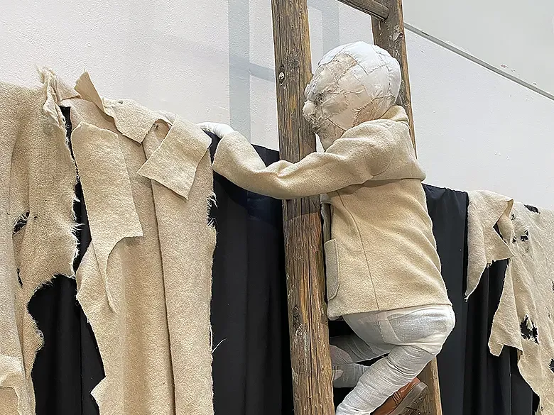 Boy with creamy coloured jacket on ladder climbing up a black curtain with shredded bright fabrics.