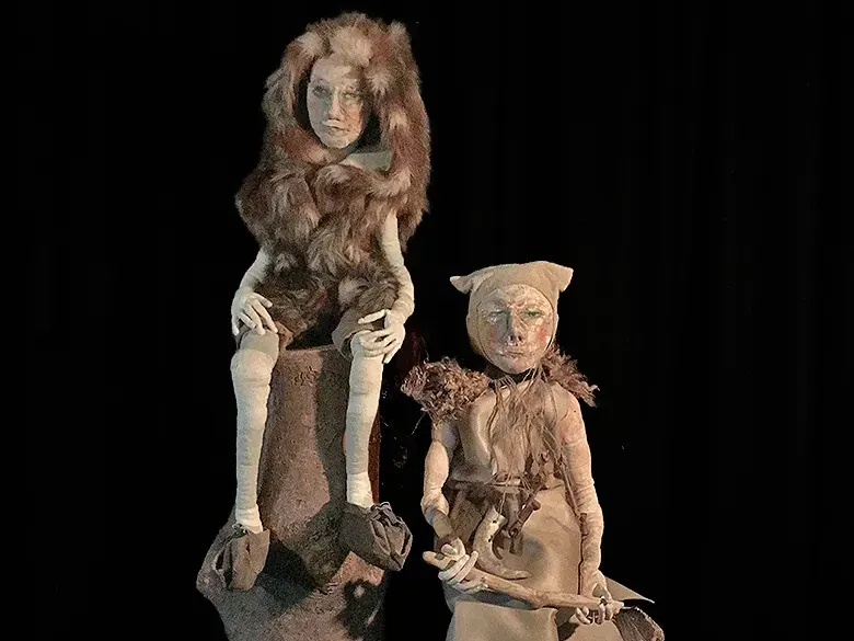 Two male sculptures in prehistoric clothes in front of dark background.
