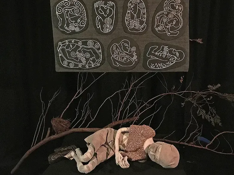 Boy sculpture lying on dark ground in front of a branch and ornamental stitchwork.