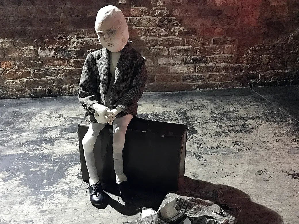 A boy sculpture wearing a tweed jacket sitting on a suitcase with shed fabrics on the floor.