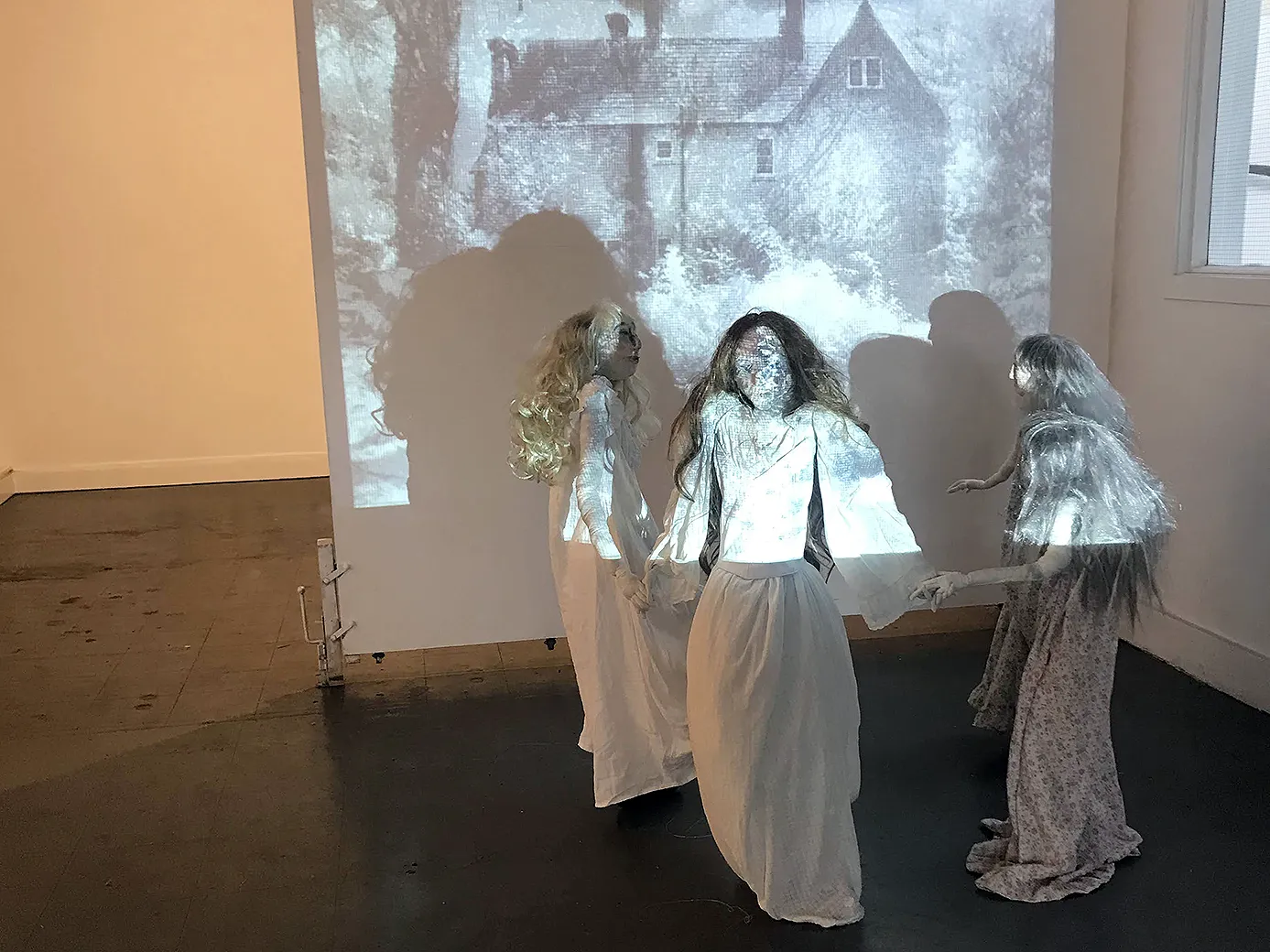 Four girl sculptures in white spooky dresses holding hands and dancing in front of black and white house projection.