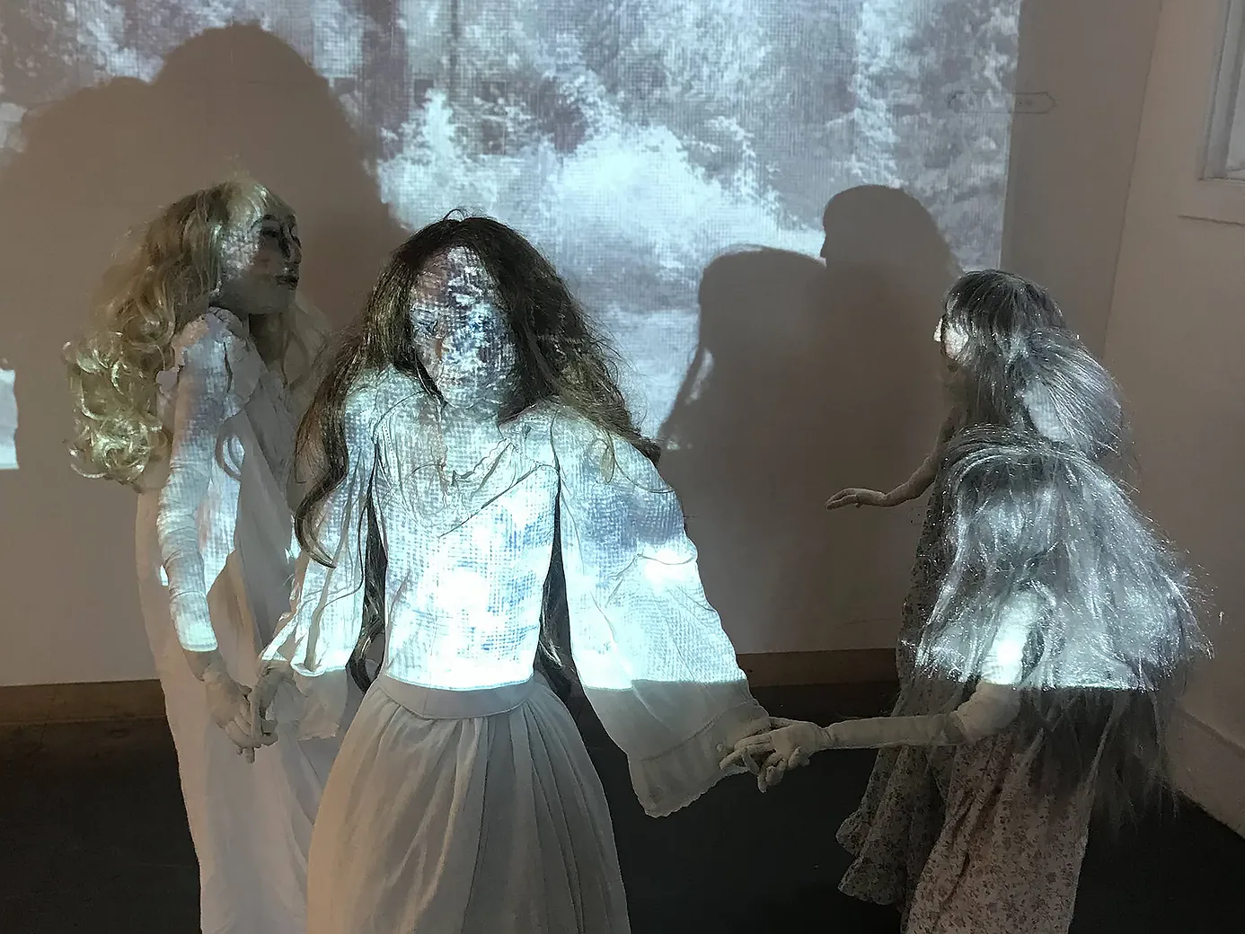 Four girl sculptures in white spooky dresses holding hands and dancing in front of black and white projection.