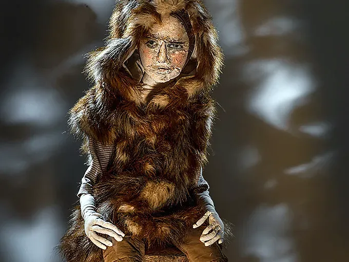 Boy sculpture in fur coat sitting on a tree trunk before a dark background.